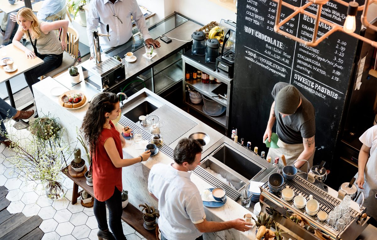 A busy cafe seen from above, with customers at the counter, staff behind a coffee machine and two stainless sinks in the bench