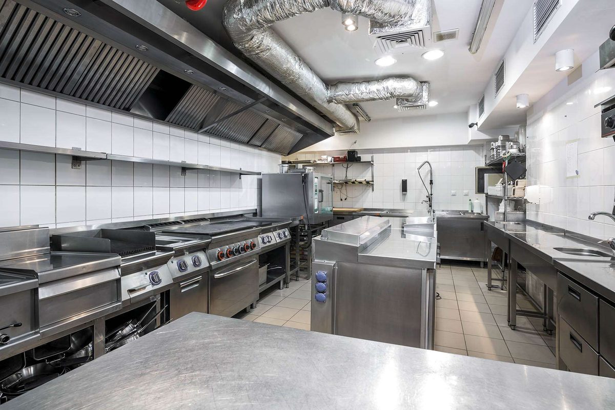 A commercial kitchen with stainless steel benches, cooking ranges, a long extraction hood and white tiled walls