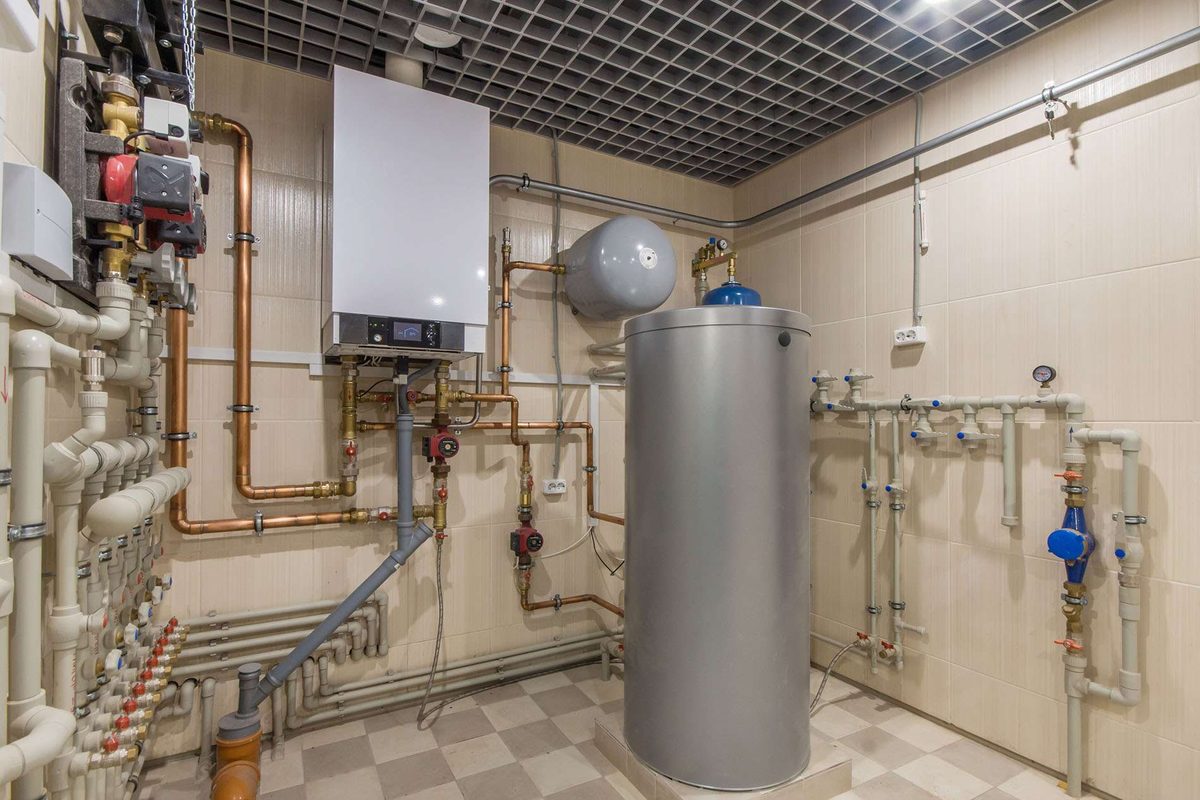 A tiled plant room with a tall silver hot water cylinder, a wall-mounted heater and copper and plastic pipework with valves and gauges