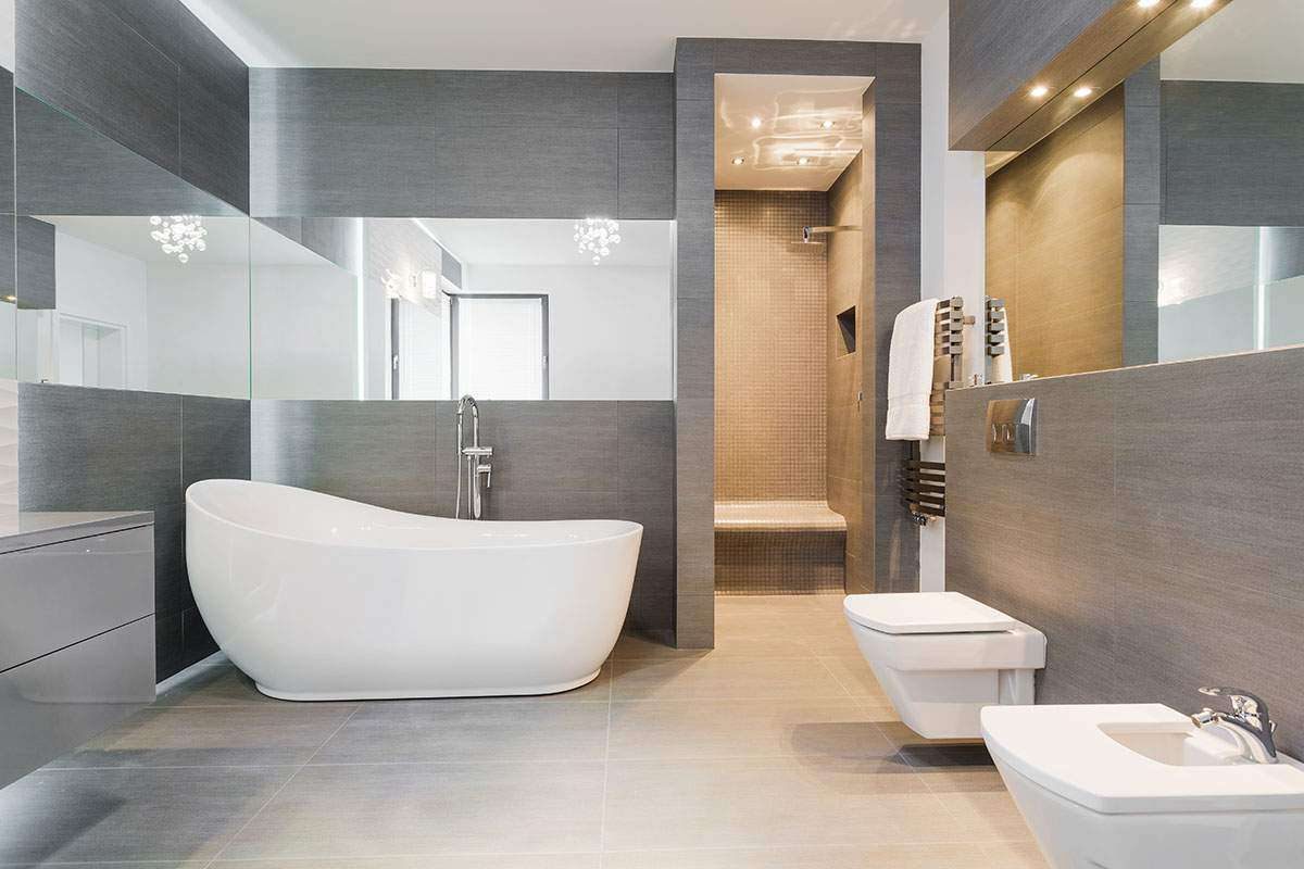 A grey tiled bathroom with a white freestanding bath, a floor-mounted bath tap, a wall-hung toilet and a walk-in shower