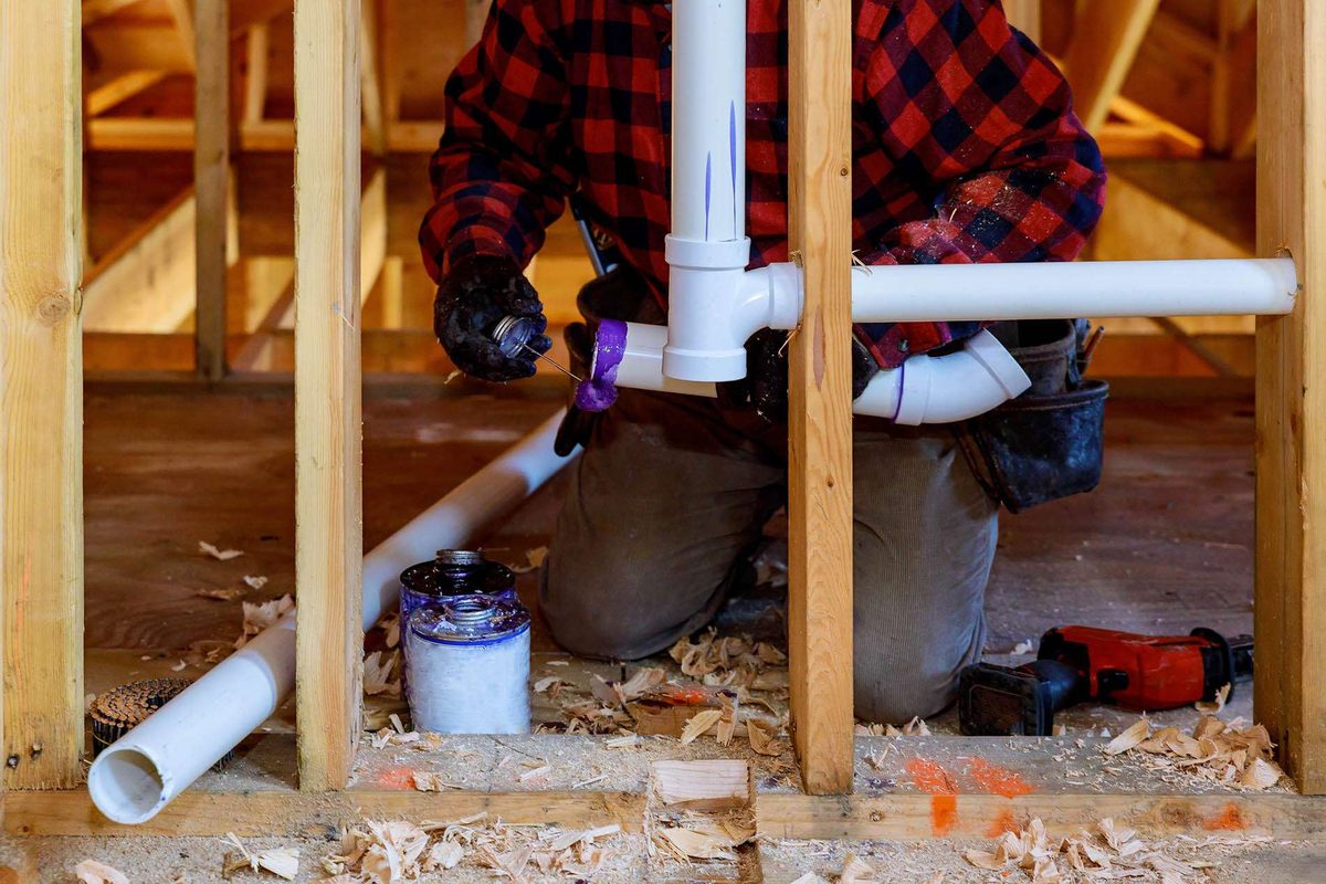 A plumber in a checked shirt and gloves kneeling inside new timber framing, joining white plastic waste pipes
