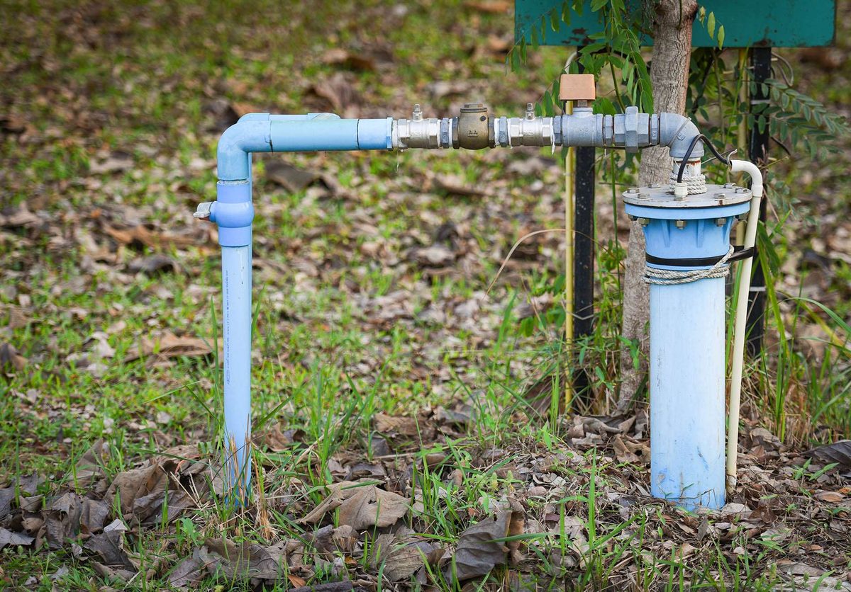Pale blue water pipework and a fitting rising out of the ground beside a pump housing, among grass and fallen leaves