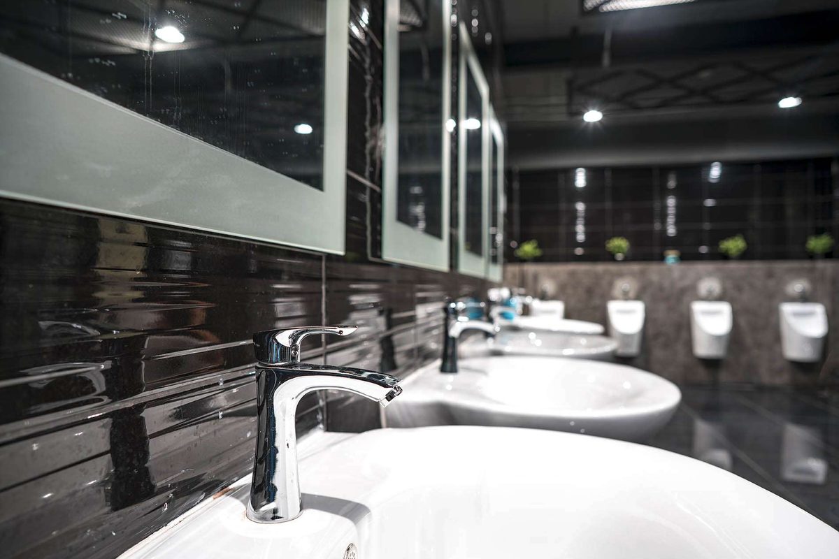 A commercial washroom with a row of white basins and chrome mixer taps on black tiles, and urinals at the far end