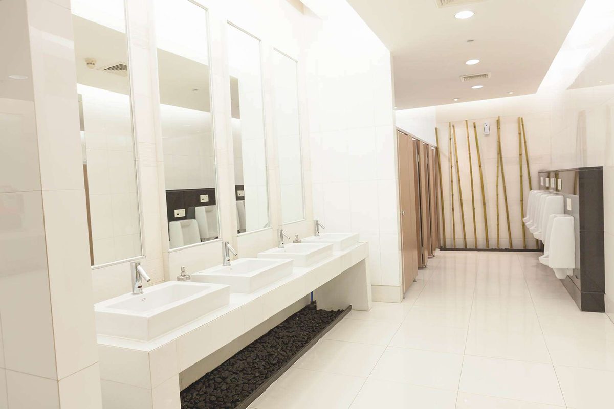 A bright commercial washroom with a row of white basins under tall mirrors and urinals along the far wall