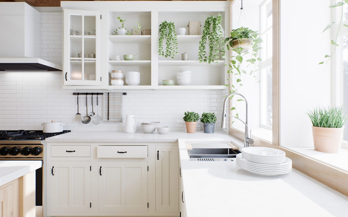 A white country-style kitchen with open shelves of plants and crockery, a gas cooktop and a sink under a sunny window