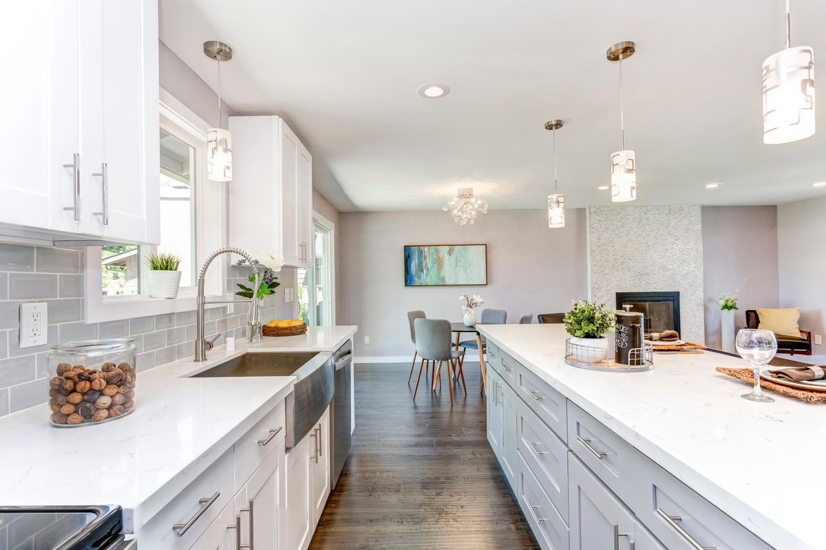 A long white kitchen with grey subway tiles, a farmhouse sink, a white stone island and pendant lights, opening onto a dining area
