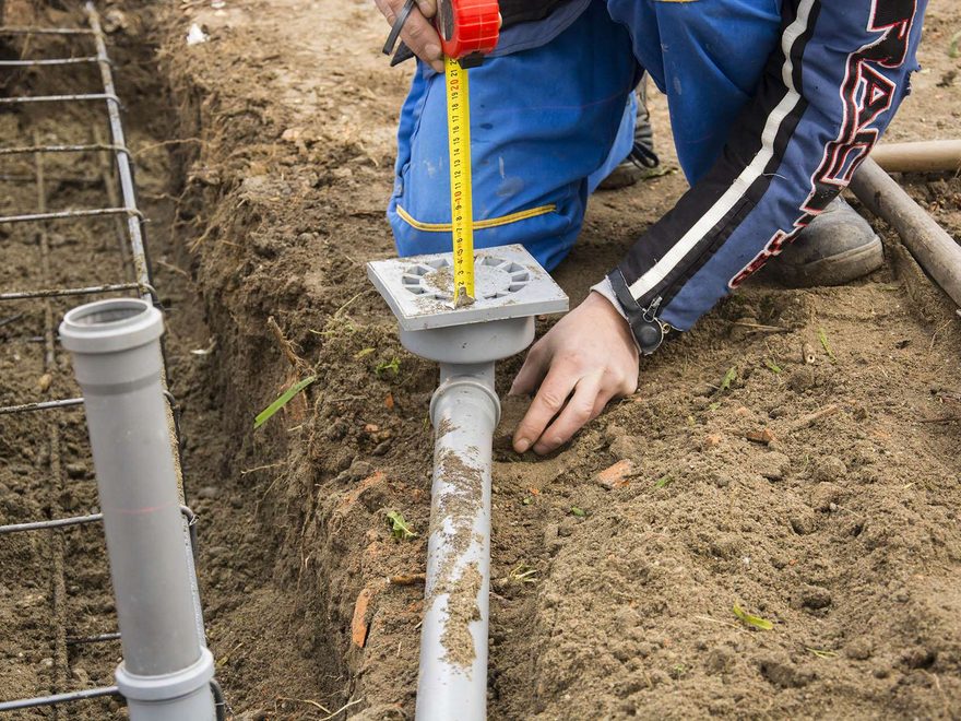 A worker crouched in a dirt trench measuring the height of a grey drain pipe and floor waste with a tape measure