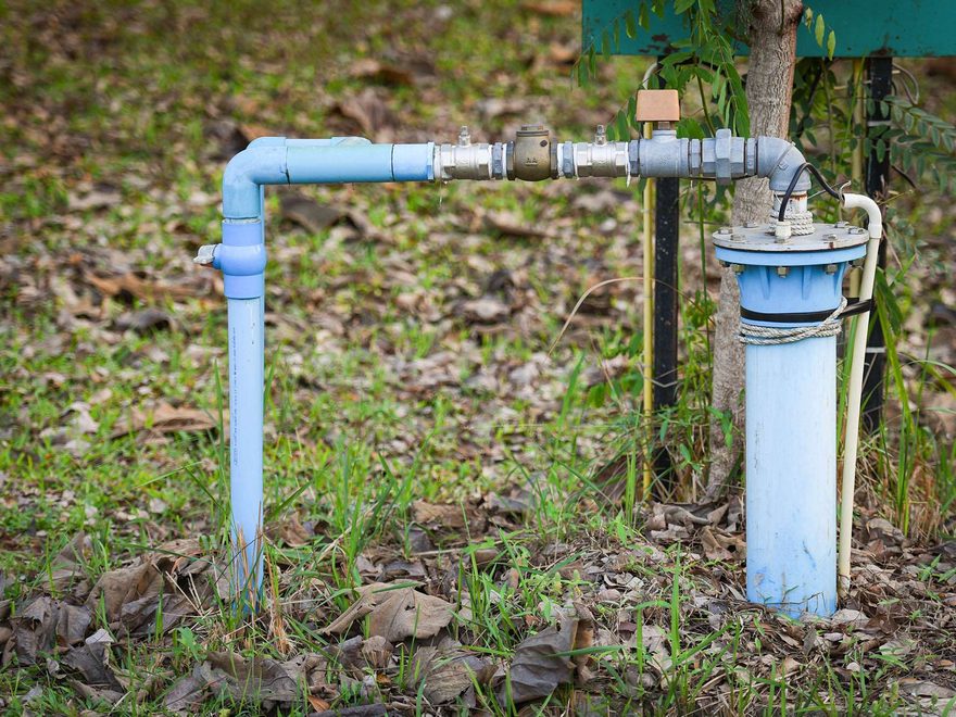 Pale blue water pipework and a fitting rising out of the ground beside a pump housing, among grass and fallen leaves
